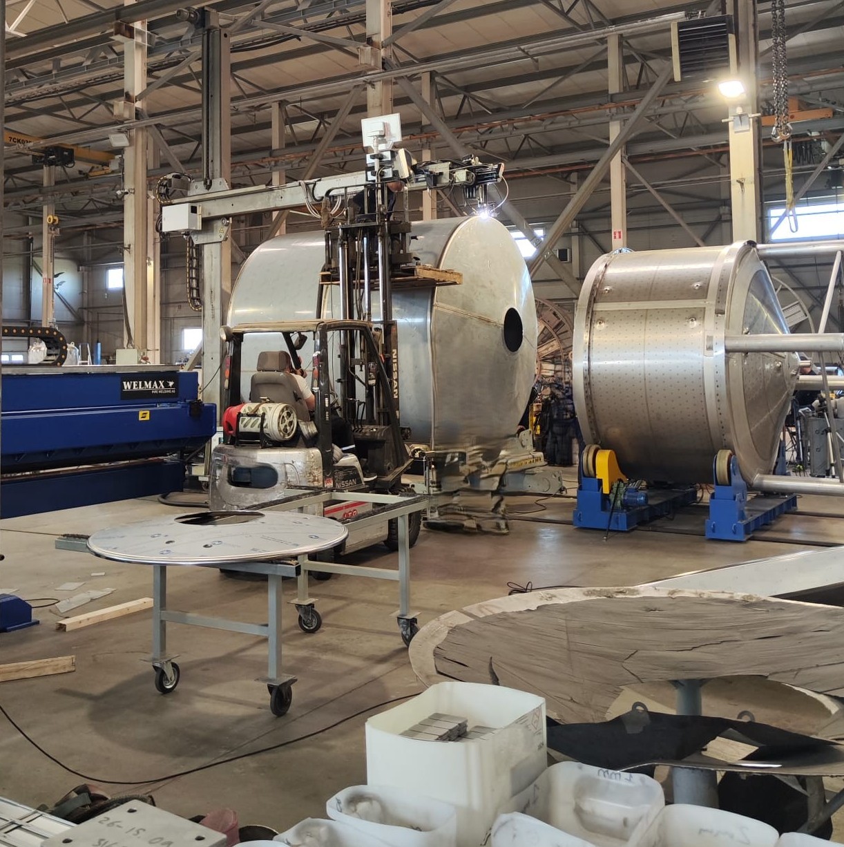 Industrial workshop with large cylindrical metal tanks, robotic welding equipment, and overhead crane inside.